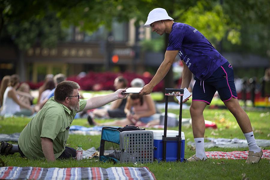 An economics student wearing a purple shirt and white hat hands a survey to someone sitting in a lawn chair at a park.